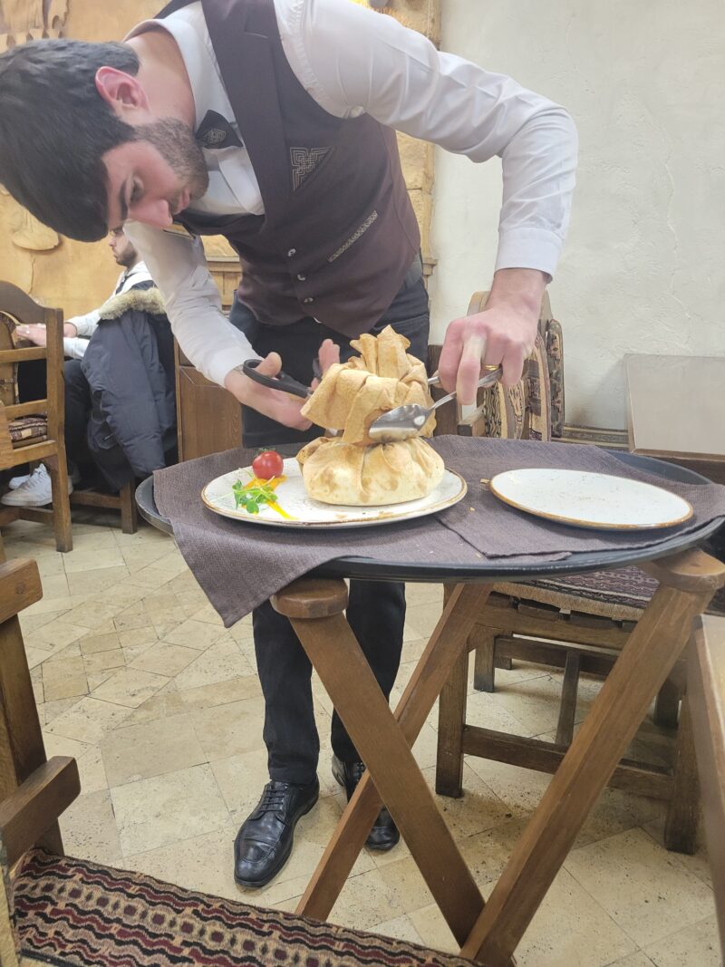 Waiter Preparing Traditional Dish – A waiter in a vest and bowtie carefully cuts open a crispy pastry dome at a restaurant, revealing the dish inside. The setting is cozy with wooden furniture and rustic decor.