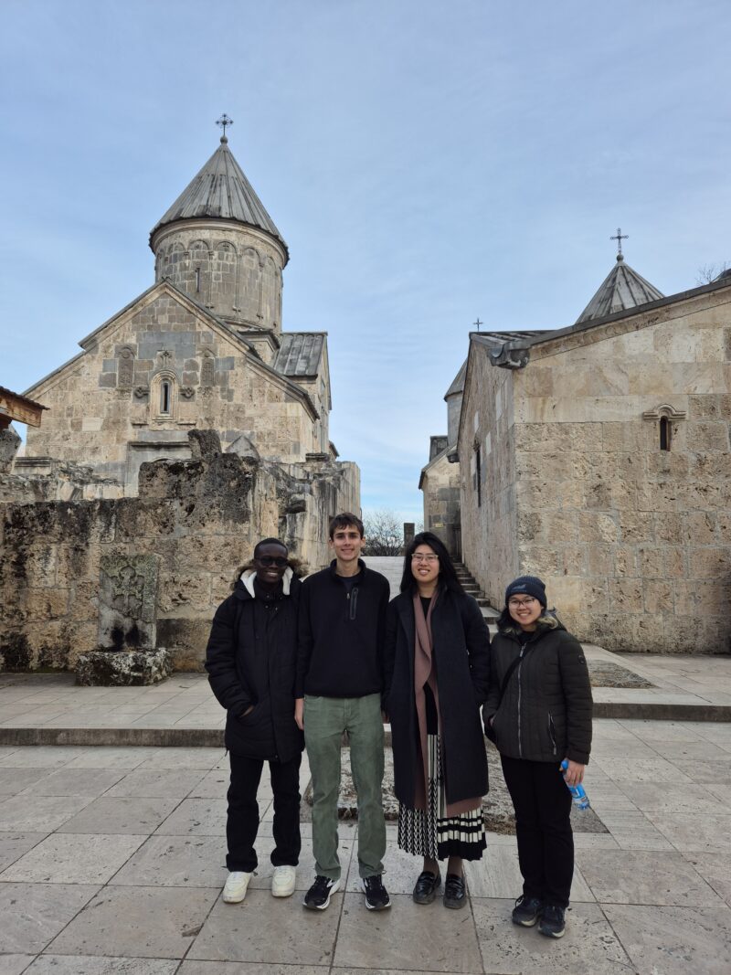 Group Photo at Haghartsin Monastery – Four friends stand in front of the ancient stone Haghartsin Monastery in Armenia, smiling on a crisp, sunny day. The monastery’s distinctive conical dome and intricate stonework rise behind them.
