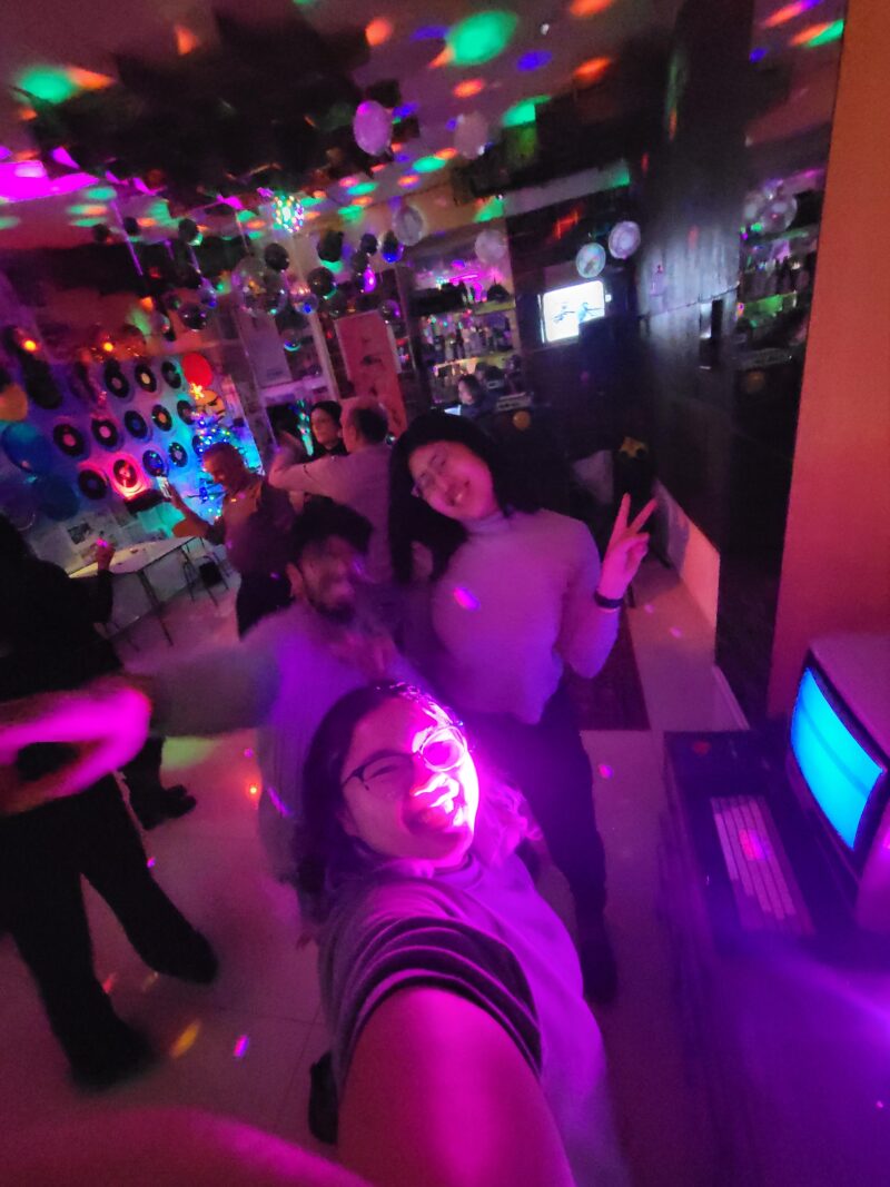 Vibrant Disco Party Selfie – A group of people dance under colorful disco lights, with one person taking a bright pink-lit selfie. The background features vinyl records, decorations, and a retro computer screen.