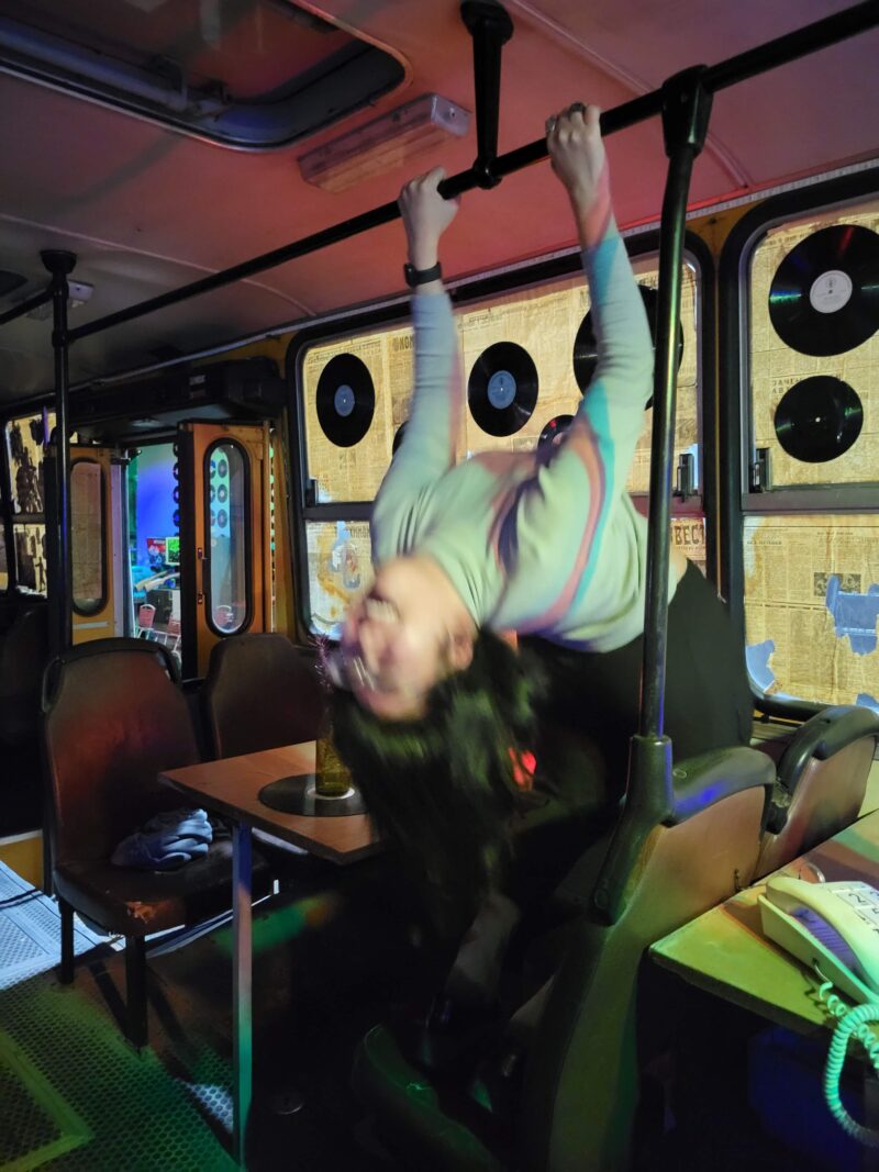 Hanging from a Bus Rail in a Retro-Themed Bar – A person in a light-colored sweater hangs upside down from a metal rail inside a bus-themed bar, surrounded by vintage seats, records on the walls, and colorful lighting.