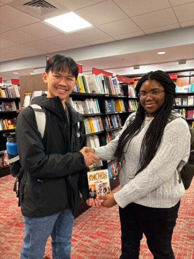 A boy and a girl (Christine) holding a One Piece book in a bookstore. They are smiling and using their other free hands to shake hands