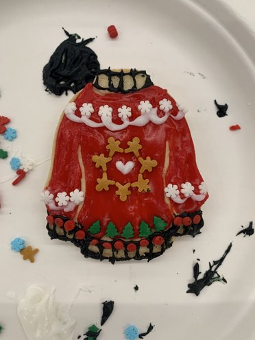 A decorated red cookie shaped like a sweater, featuring white icing, green trees, and festive detailing.