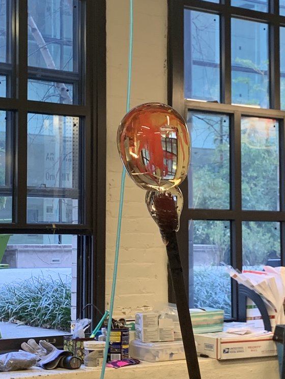 A close-up of a glassblowing tool with a molten glass piece being shaped, against a backdrop of large windows and studio materials.