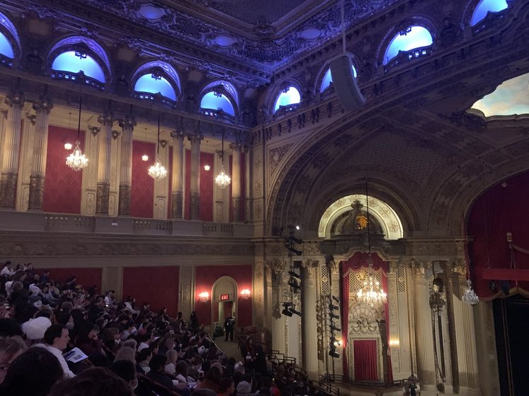 An ornate theater filled with an audience, showcasing grand chandeliers and decorative architecture under soft, blue lighting.