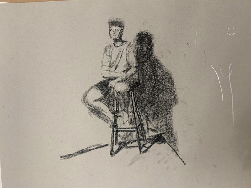 The sketch depicts a male figure rendered in charcoal on textured grey paper. The figure is seated on a simple wooden stool, with a relaxed posture, arms resting on his knees. The light source appears to be above and slightly to the right, creating a pronounced shadow to the left of the figure. The shadow emphasizes his shape and adds dimensionality to the composition, while faint texture marks on the background, possibly from the drawing process, are visible.