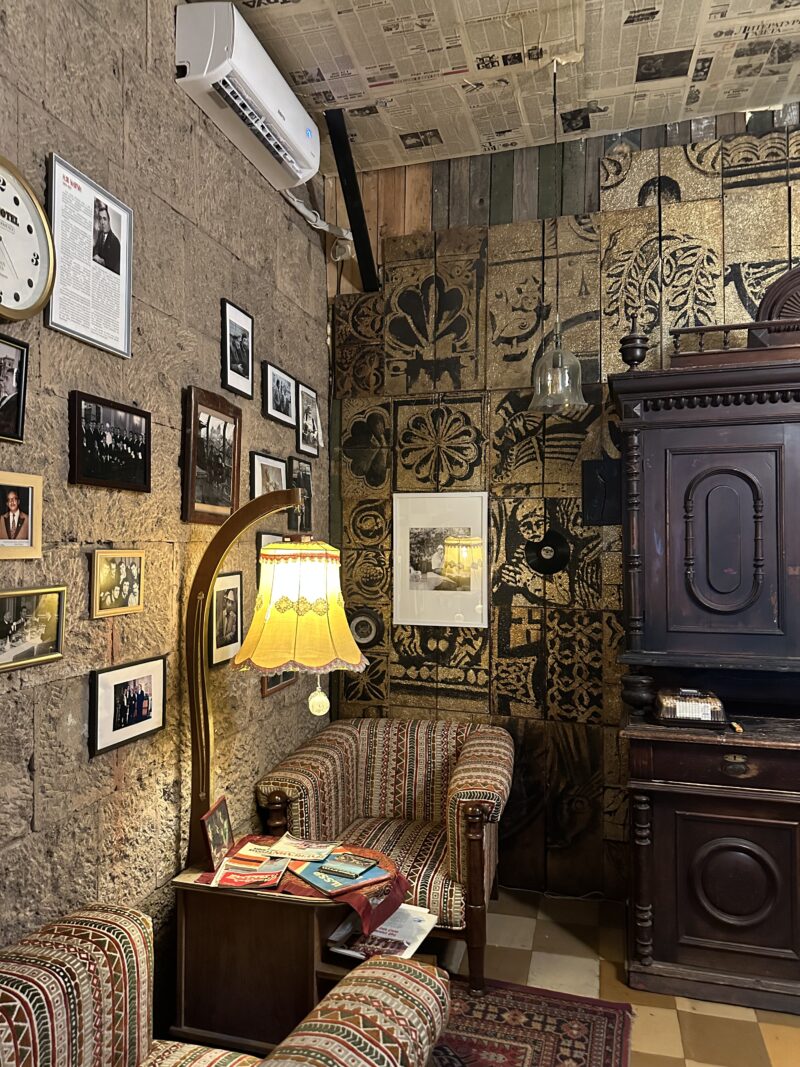 Cozy Vintage-Themed Lounge – A warmly lit corner of a lounge features patterned armchairs, framed black-and-white photos, a yellow lampshade, and walls covered with old newspapers and golden patterns.