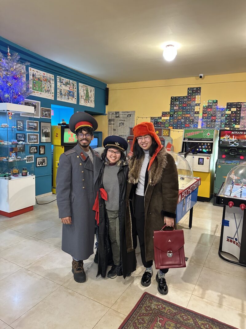 Dressed in Soviet-Era Military Coats and Hats – Three friends pose in a vintage arcade museum wearing oversized Soviet-era military coats and hats, standing among old-school games and memorabilia.