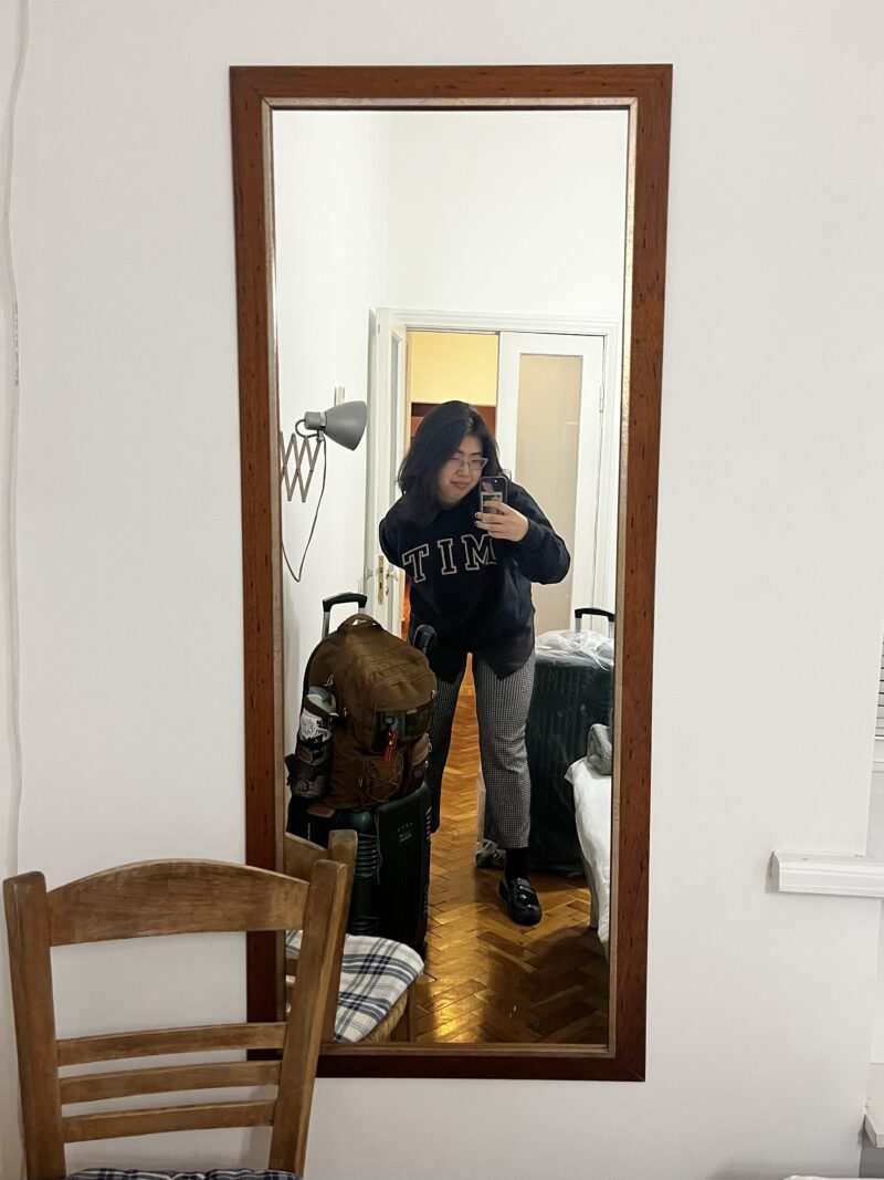 Mirror Selfie in a Room – A person wearing a black MIT sweatshirt and glasses takes a mirror selfie in a warmly lit room. Luggage, a wooden chair, and a backpack are visible in the background.