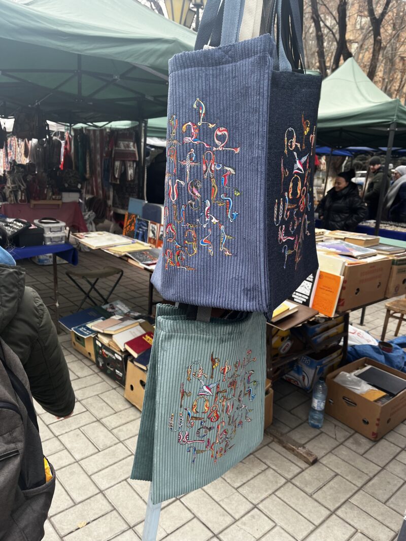 Market Stall with Embroidered Tote Bags – Two handmade tote bags with intricate, colorful embroidery hang from a stall at an outdoor market. Other handmade crafts, books, and fabrics are displayed in the background.