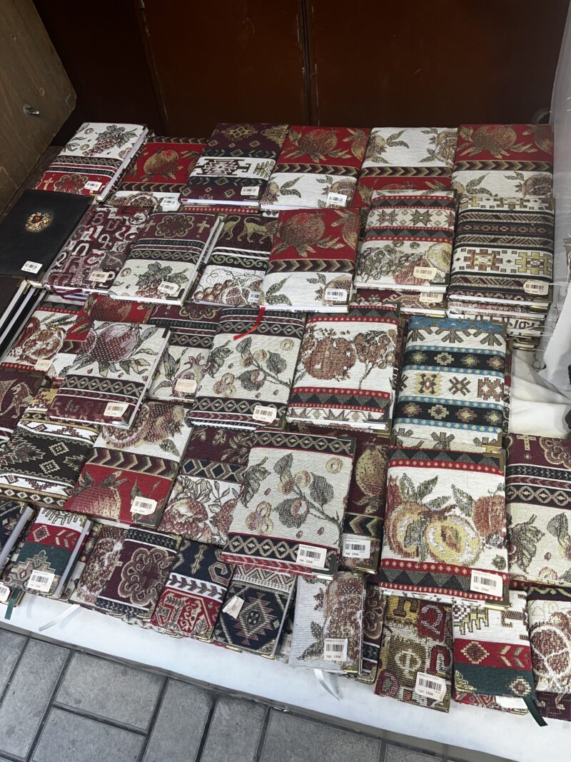 Tapestry Notebooks at a Market Stall – A table full of richly patterned notebooks with tapestry-like fabric covers, featuring traditional motifs, is arranged neatly at an outdoor market.
