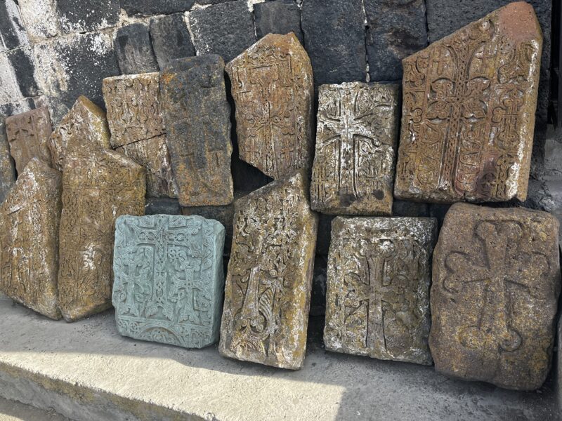 Ancient Khachkars (Cross-Stones) Against a Wall – A collection of Armenian khachkars, ornately carved cross-stones with intricate patterns, rests against a black stone wall. One stone is a striking blue-green, contrasting with the others' earthy hues.