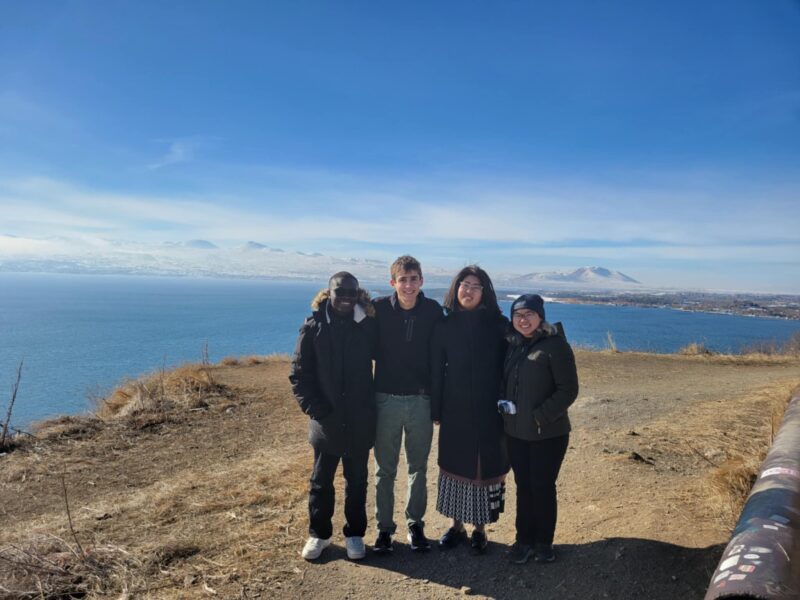 Group Photo with Lake Sevan in the Background – Four friends stand on a hilltop overlooking the deep blue waters of Lake Sevan, with snow-capped mountains stretching into the horizon under a clear sky.