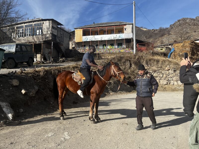 Horseback Riding in a Rural Village – A young man rides a brown horse in a rustic Armenian village while a local guide holds the reins. Behind them, traditional stone houses with colorful decorations stand under a bright blue sky.