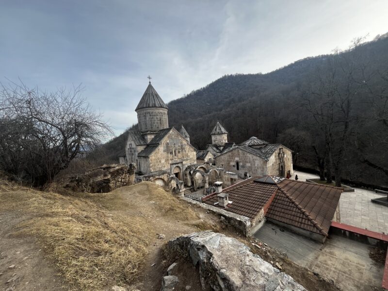 An ancient monastery nestled in the mountains, surrounded by bare winter trees and rugged terrain.