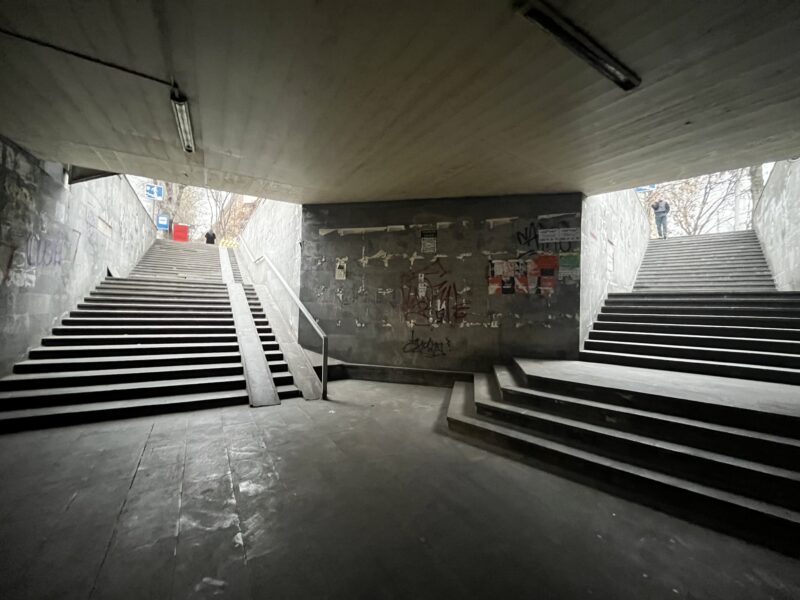 Urban Underpass with Graffiti – A dimly lit pedestrian underpass with wide concrete stairs leading up to the street. The walls are covered in posters and graffiti, and two people are seen exiting.