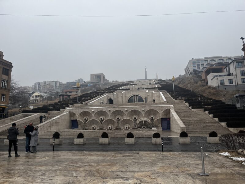 Yerevan Cascade on a Foggy Day – A wide stone stairway with cascading fountains and geometric designs ascends a hill in Yerevan, Armenia. Visitors are scattered along the steps, and modern buildings rise in the background.