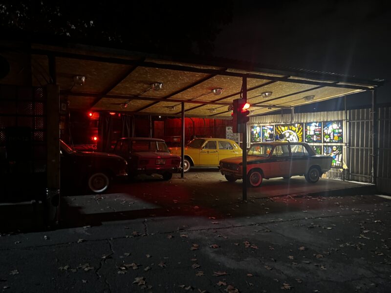 Retro Car Display with Neon Lights – A dimly lit garage houses several old Soviet-era cars, illuminated by red and yellow neon lights and a stained-glass window in the background.