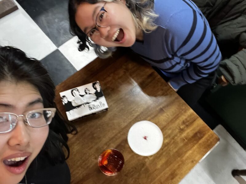 Excited Drinks and Game Night – Two friends pose with open-mouthed excitement over a table with drinks, a Beatles-themed card game, and a checkered floor in the background.