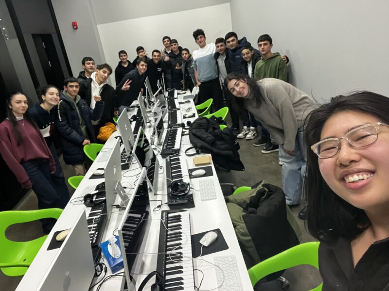 Group Photo in a Computer Lab – A large group of students and instructors pose for a selfie in a well-lit computer lab with green chairs and Mac computers. The group is smiling and making peace signs.