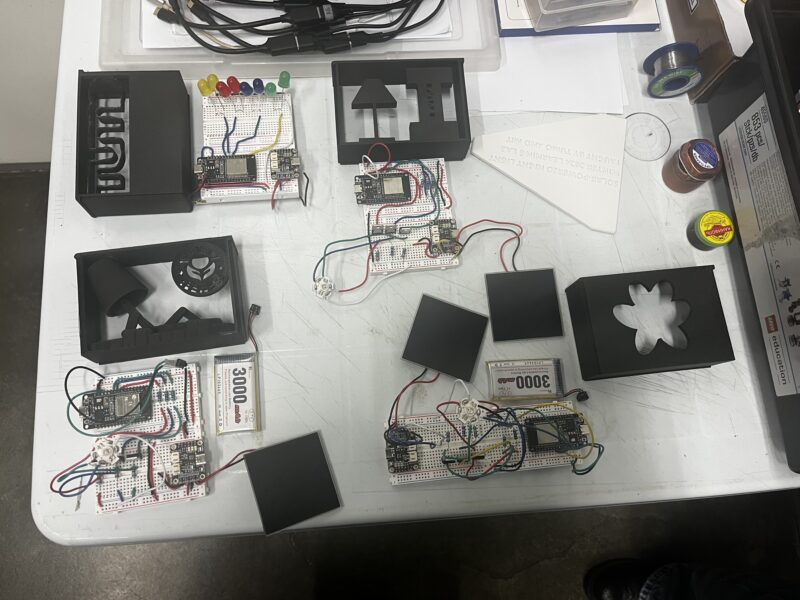 Electronics Prototypes on a Workbench – Various electronics projects with circuit boards, LEDs, batteries, and 3D-printed enclosures are laid out on a table, appearing to be part of a hands-on workshop.