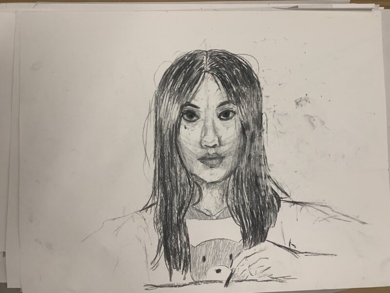 The image is a pencil sketch on white paper, showcasing a young woman with long dark hair parted in the middle. Her facial features are rendered with fine lines and shading, capturing a thoughtful expression. The composition centers around her face, with minimal background detail, emphasizing her features. The use of varied line thickness and shading creates depth, while stray pencil marks and smudges around the edges indicate the artist's process.