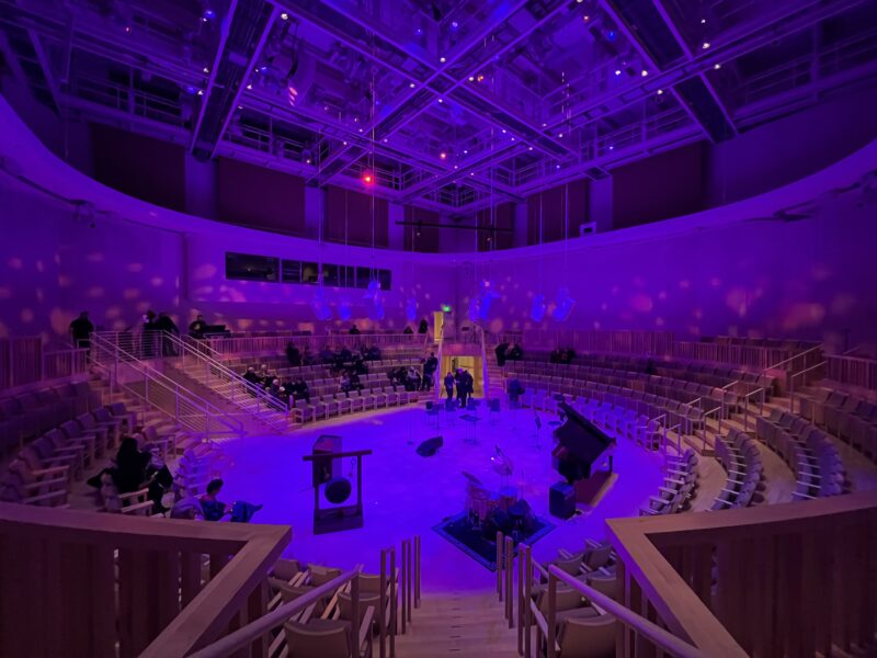 A view from the upper level of the concert hall. Purple lights envelope the space, with the stage at the center.