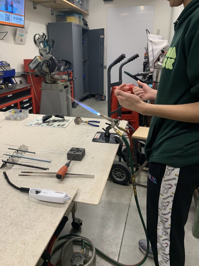 A person in a green hoodie operates a blowtorch, surrounded by various tools on a workspace in a workshop. A TV displays a timer in the background.