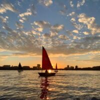 Sailboats with colorful sails glide across shimmering water at sunset, with a city skyline silhouetted…