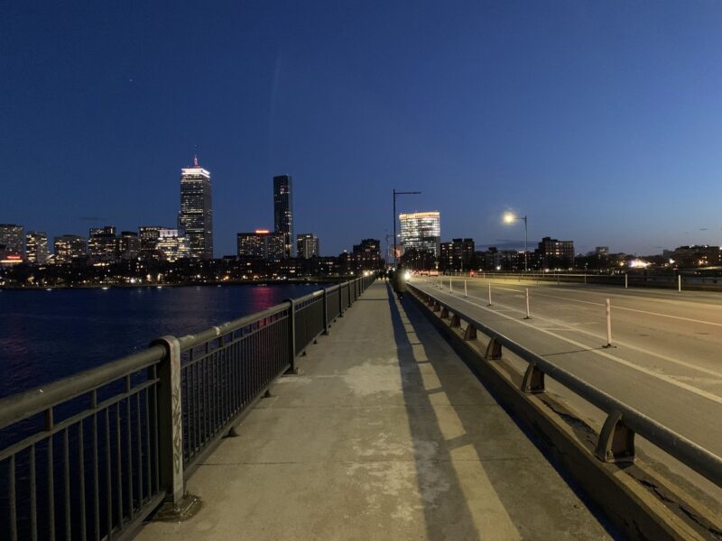 A wide view of a bridge at dusk, with city skyline lights reflecting on the water and a clear blue sky overhead.