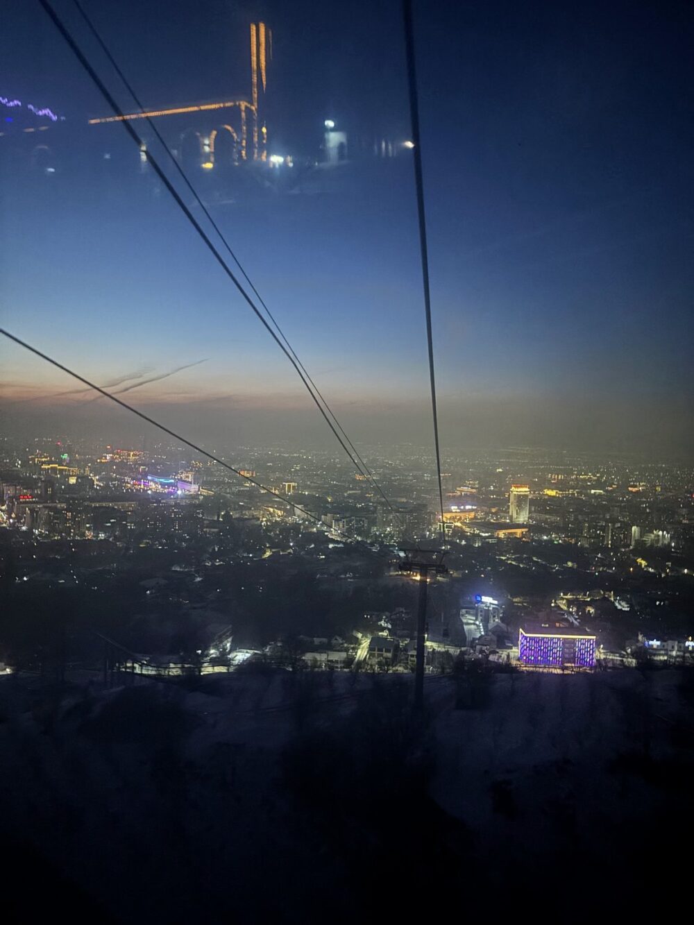 Wide shot of a cityscape from a high vantage point at dusk. The composition features strong leading lines from the cable car wires that intersect the horizon. Various sized and bright lights create a depth of field and focal points through the haze of the city. A warm gradation in the upper sky contrasts with the cooler, deeper blue of the night sky. The foreground is underexposed, providing a sense of height and scale.