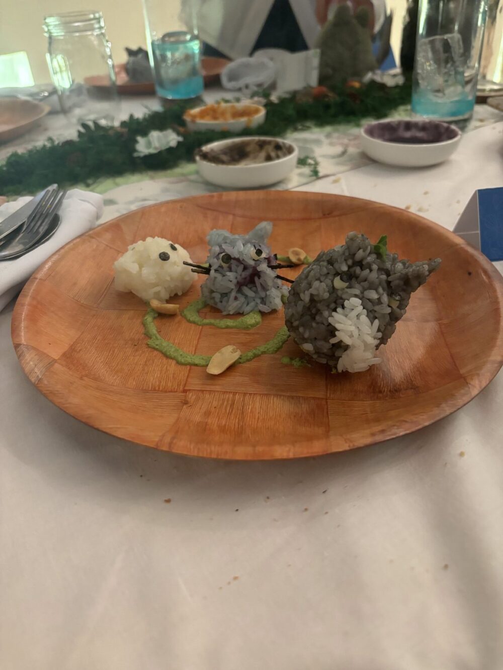 Medium shot of a wooden plate on a linen tablecloth. The plate contains three anthropomorphic rice ball formations, garnished with green sauce and nut slivers. The background is blurred with several bowls and cups. The lighting is slightly diffused. Predominant colors : muted greens, various shades of grey, beige. The composition focuses on the rice ball creatures, inviting the viewer to observe their intricate detail.