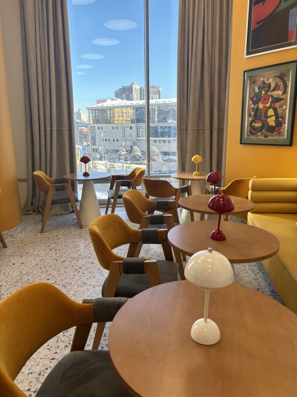 A high-angle, interior shot showcases a cafe space. The color palette centers on golden yellows and natural wood tones. The foreground features several round tables arranged with coordinating chairs. Lighting includes natural daylight from a large window on the left, and three table lamps, each on a different table, in red, yellow, and white. In the background, the cityscape is visible through the window. The right wall is a solid yellow with colorful contemporary artwork hanging on it. Composition highlights the furniture arrangement and the visual interplay between indoor and outdoor elements.