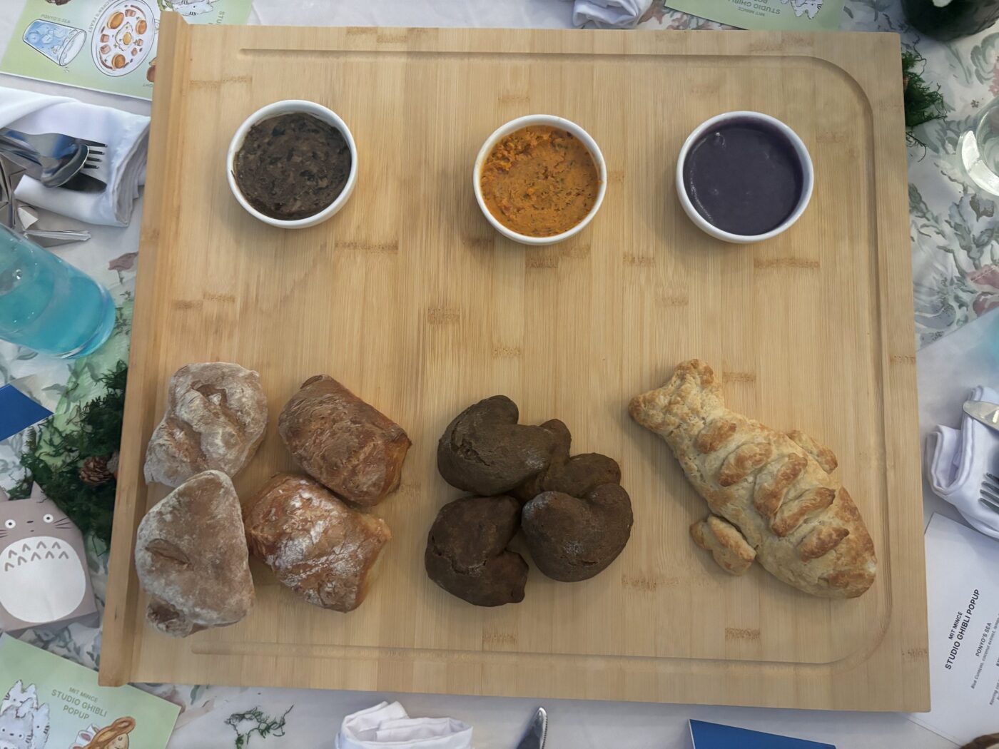 Overhead shot of a rectangular wooden cutting board featuring a row of various artisan breads; rustic dark breads, and a pastry baked to resemble a fish. Above the bread are bowls filled with a purple, orange, and brown sauces. The edges of the board intersect the corners of the frame diagonally, resulting in a high-angle composition. The event appears to be Studio Ghibli themed due to the menu text and character drawings on the table around the board.