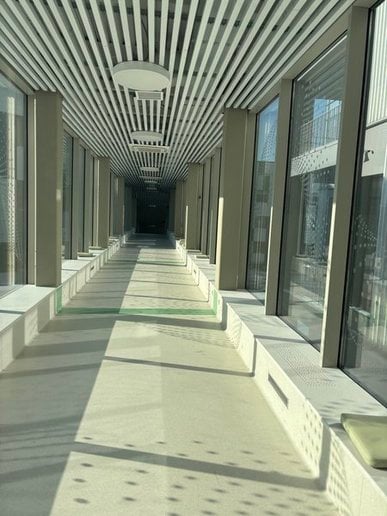 A high-angle, wide shot of a hallway. The ceiling is comprised of white slats with evenly spaced circular lights. The floor is light beige, with the hallway disappearing into darkness in the distance. Tall support beams, and large windows run along both sides of the corridor. Strong sunlight penetrates through the windows creating sharp shadows. The architectural design suggests a modern or contemporary style.