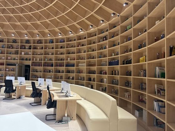 Wide-angle shot of a library interior. Dominant vertical lines of the light wooden shelves, horizontally divided and filled with diverse objects. Foreground includes light wooden desks with desktop computers and black office chairs, placed along a curved, light beige bench. The ceiling is a complex wooden grid pattern with recessed lighting. Color palette is muted with light wood tones contrasted by the black chairs. Lighting is soft and diffused showcasing the depth of field.