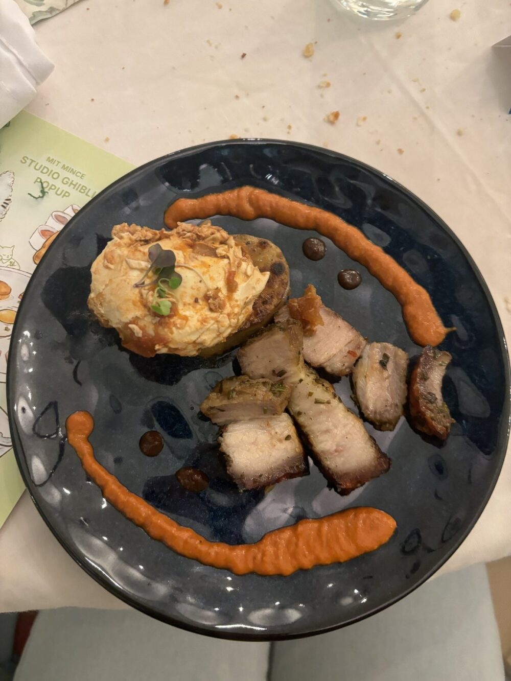 High-angle close-up shot of a black plate centered against a white table cloth. The plate features chunks of pork belly and a potato cake topped with creamy sauce. A bright orange sauce surrounds the plate, while a umber-colored sauce spot the plate among the sauces. In the left of the frame, the corner of Studio Ghibli popup menu is visible. Shallow depth of field, natural lighting.