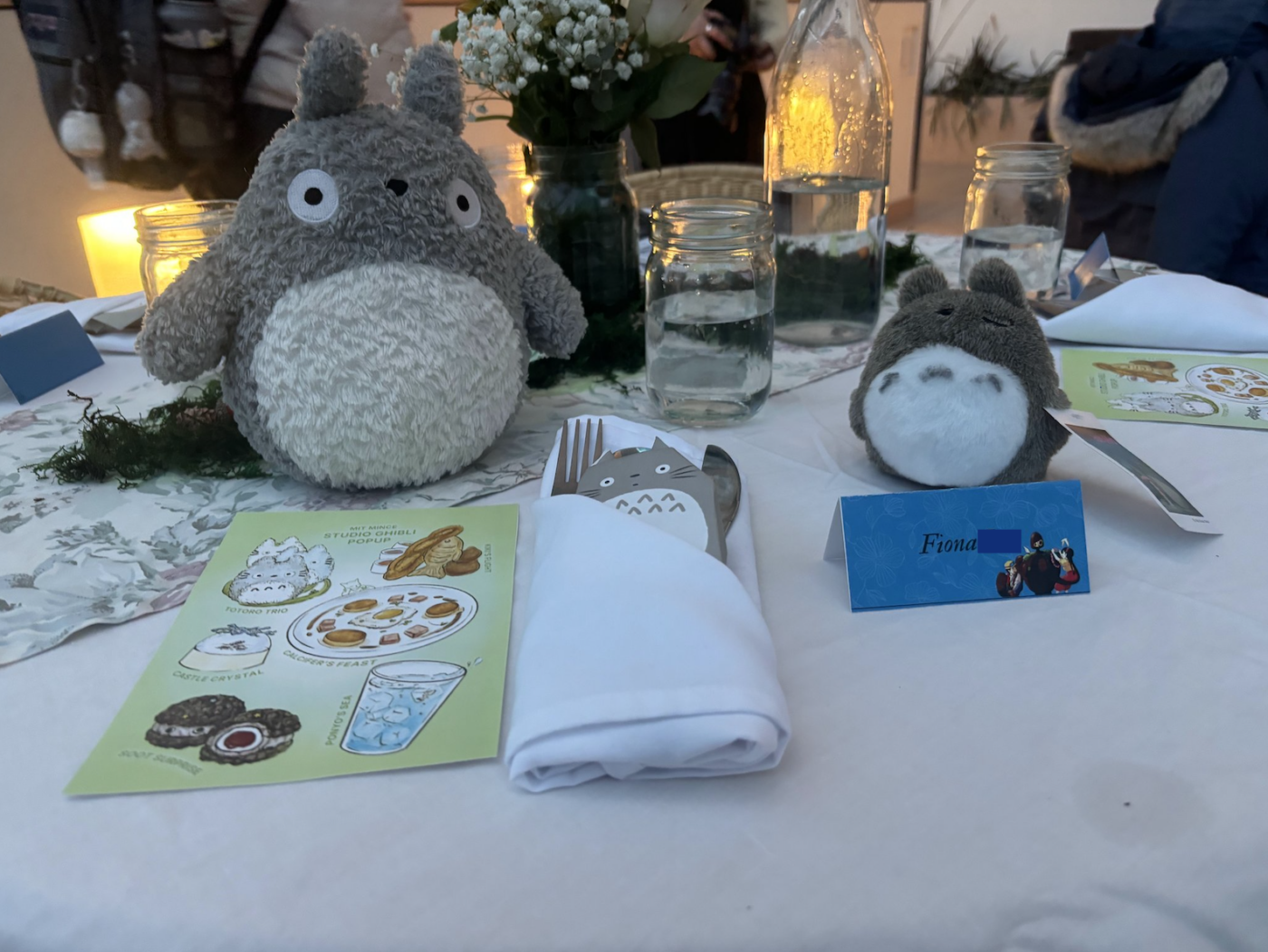 Close-up, slightly high-angle shot of a Studio Ghibli-themed table setting. A large, roughly textured, gray Totoro plushie dominates the left of the frame. It sits next to a vertically oriented, light green menu card featuring sketched dishes, with Baby's breath flowers in a vase in the background. A fork and knife are wrapped in a white cloth containing a paper Totoro detail. Positioned on the right is a smaller Totoro plushie with a blue place card titled "Fiona." Each setting is carefully positioned on a wrinkled white tablecloth under soft, warm lighting.