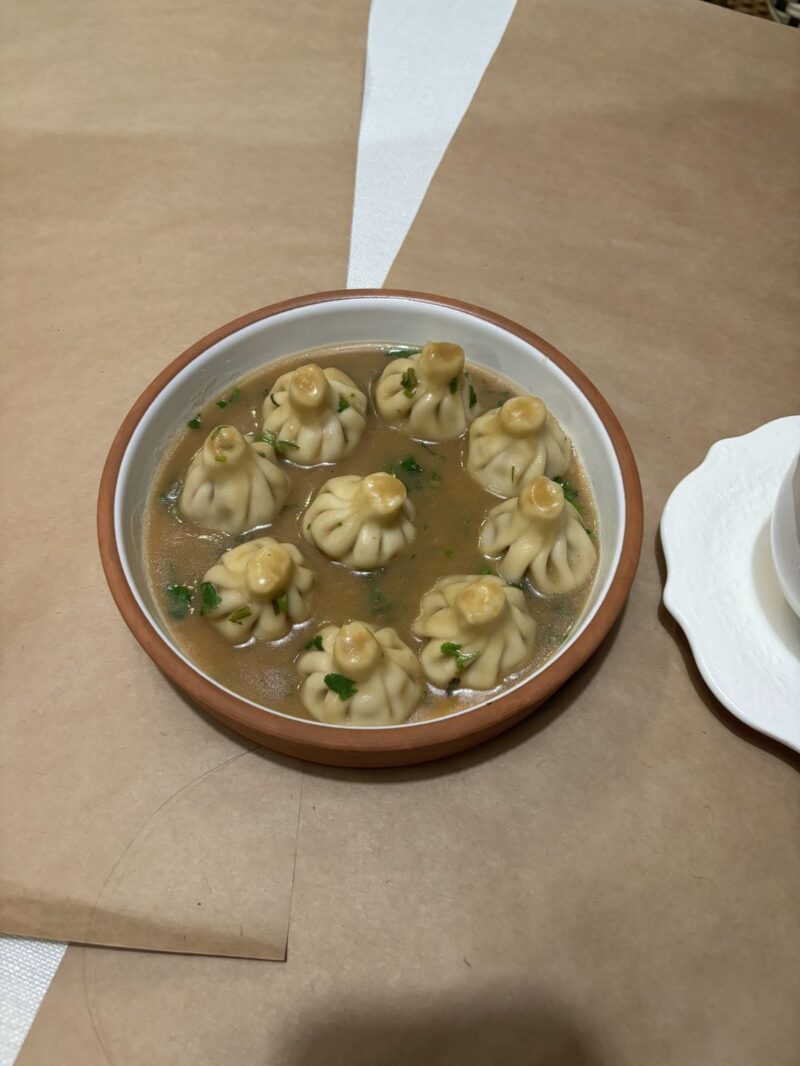 High-angle, medium shot of a round earthenware bowl with a white interior containing seven khinkali dumplings, submerged in a light brown broth and garnished with green herbs. The bowl is placed against a brown kraft paper background, with a white, decorative plate partially visible on the right side. Lighting is soft, highlighting textures of the dumplings and the bowl.