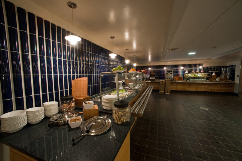 Looking down a kitchen of a dining hall, with black floors and a long black countertop filled with food going down and around the back the of the room.