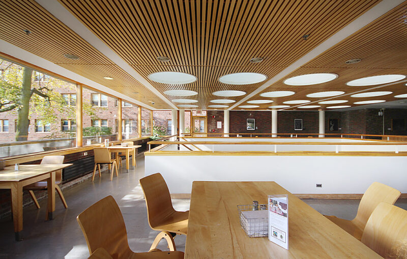 A dining area with natural lighting and lots of windows. Tables and chairs go around the edge of the room, with the staircase going down in the middle of the room.