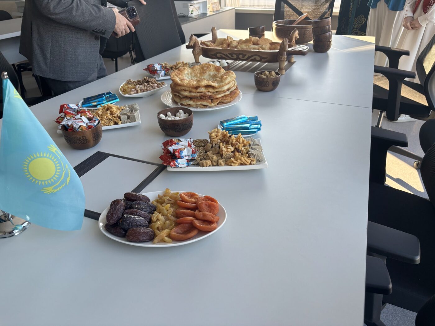 Wide-angle shot of a gray office conference table. The composition is centered on the table, which features multiple plates and bowls filled with food laid out on a table. A small Kazakhstan flag is placed in the lower-left corner. Other Kazakh cultural items and traditional clothing are visible. Soft window light is present on the outside. A portion of a man is seen on the left taking a photo of the table with his phone. The background is slightly blurred to highlight the food.