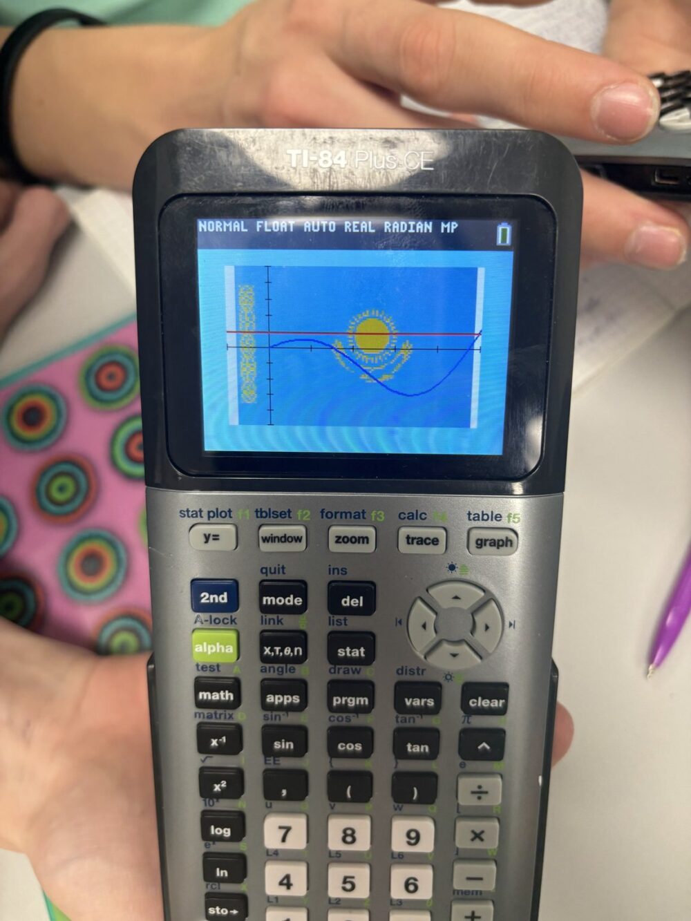 Close-up shot focuses on a TI-84 Plus CE calculator held in someone's hands. The LCD screen prominently displays a graphed sine wave (blue) and a horizontal line (red) superimposed over the Kazakh flag (light blue background with a stylized sun and eagle). The calculator's keypad is partially visible, silver and with black buttons labeled with mathematical functions. Soft, diffused lighting with shallow depth of field; elements of a classroom environment (hands, notebook) are blurred in the background.