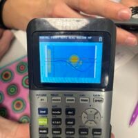 Close-up shot focuses on a TI-84 Plus CE calculator held in someone's hands. The LCD…