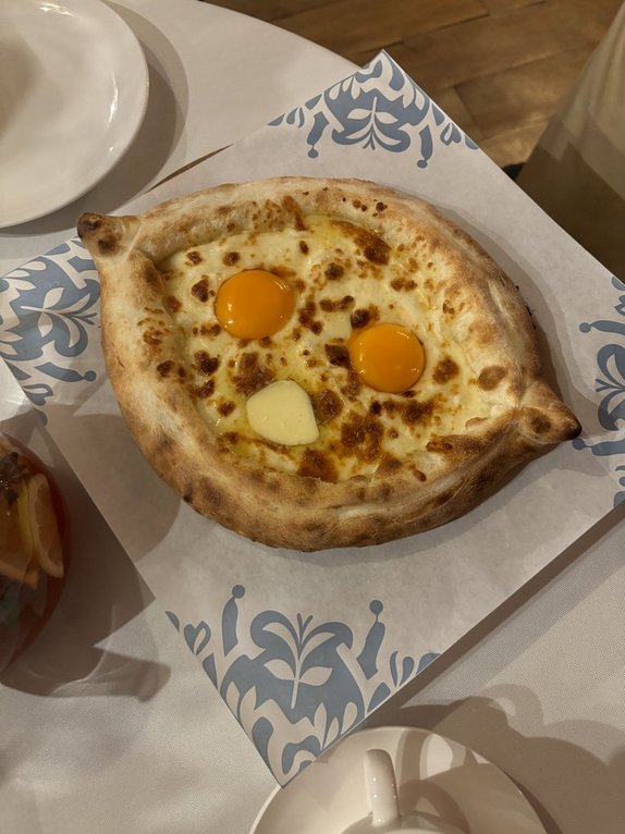 High-angle, medium shot of Khachapuri Adjaruli. The cheese boat is centered on a square, blue-patterned paper. Hints of a tea cup, small white plate, and a sangria-like beverage occupy the foreground. The lighting is soft, highlighting the golden crust of the bread and vibrant egg yolks.