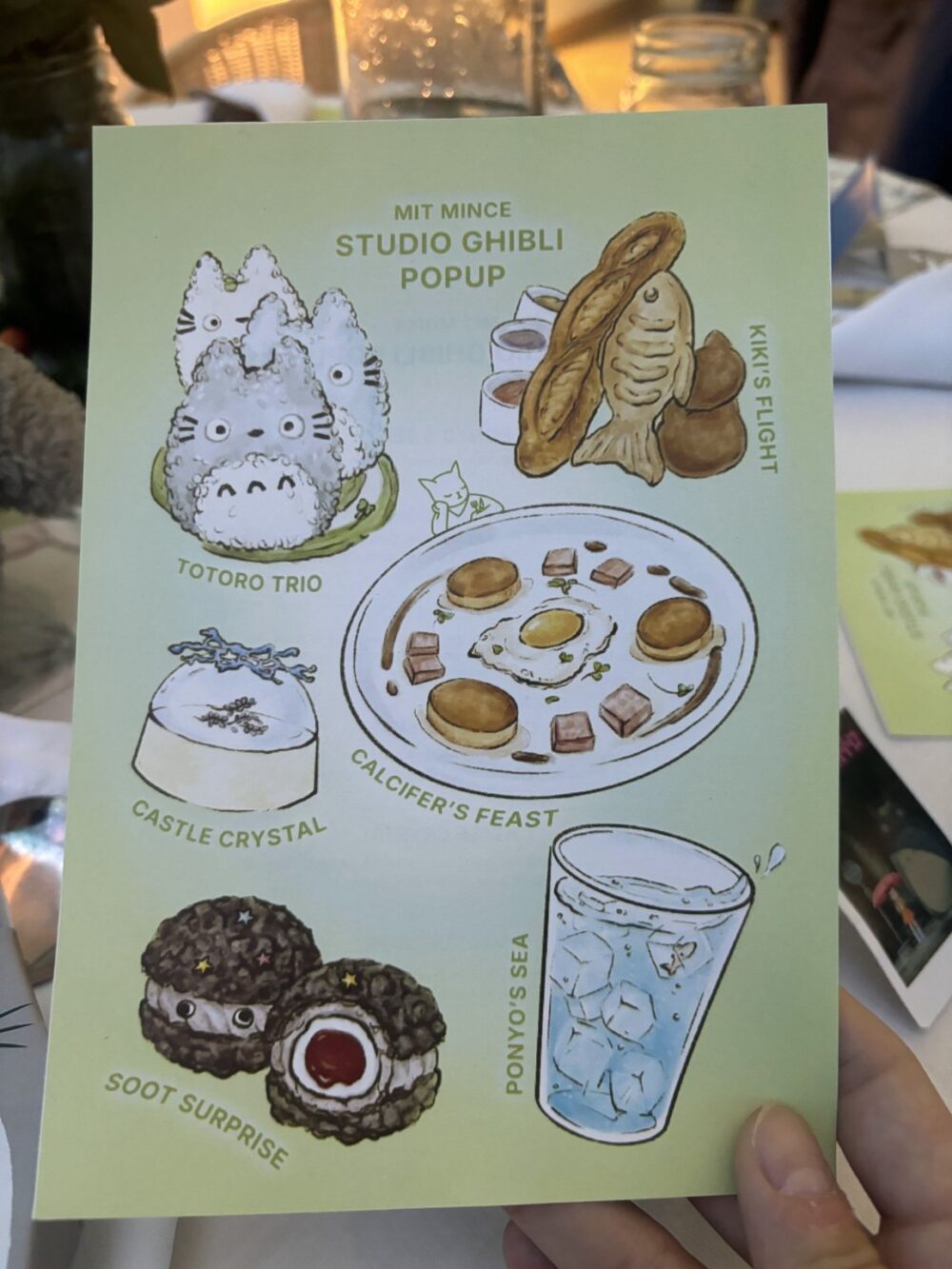 A hand-held, light green menu card features color illustrations of themed food from Studio Ghibli films. The illustrations, starting from the top-left and going down and then right include: three Totoro characters on a green bed, a baguette and brown fish, a white pudding with blue decoration, a breakfast styled food plate, two brown cookies with a red center and a glass of ice water. Text labels each food creation. The text overlays the images and says: TOTORO TRIO, KIKI’S FLIGHT, CASTLE CRYSTAL, CALCIFER’S FEAST, SOOT SURPRISE, and PONYO'S SEA. The Menu is titled: MIT MINCE STUDIO GHIBLI POPUP. The Background is blurred with various objects and colors.