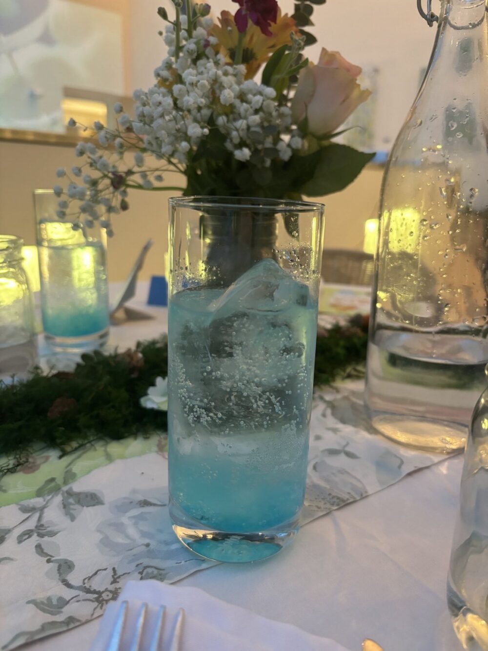 Close-up shot of a clear, cylindrical glass with a blue liquid and ice, set on a tablecloth with floral patterned. Flower arrangement in the background in a steel vase. A transparent glass bottle on the right. Shallow depth of field blurs the background details, keeping focus on the drink. The image is warm-toned, with natural lighting.