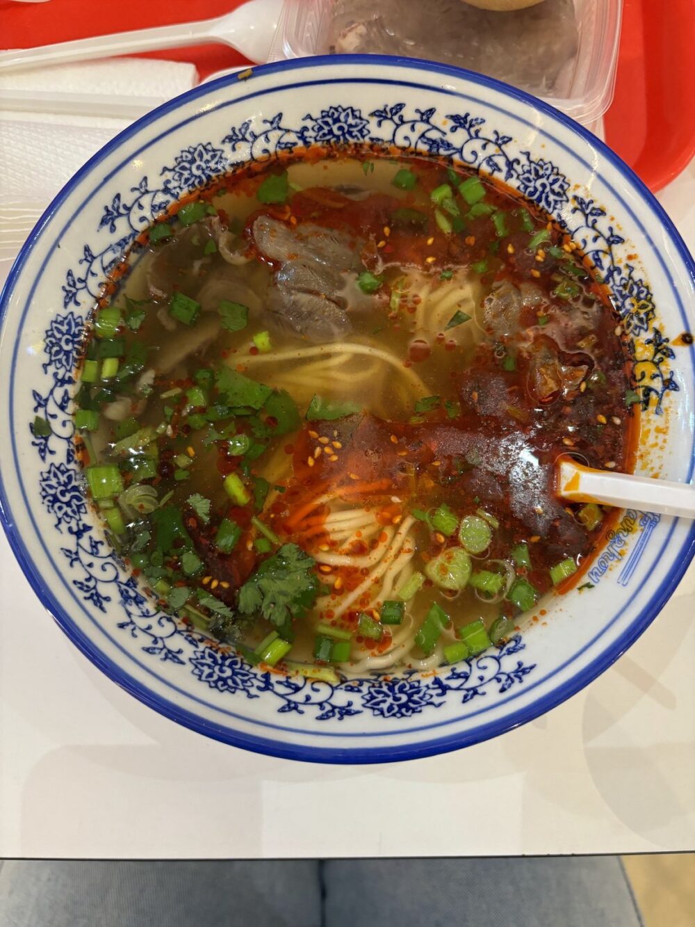 High-angle, full shot of a bowl of beef noodle soup. The bowl is round with a blue and white floral/geometric pattern around the rim. Inside, the soup contains noodles, beef slices, and chili oil, and is garnished with bright green cilantro and green onions. White chopsticks are laying diagonally across the top right edge of the bowl. The background is nondescript, and focuses attention on the food.