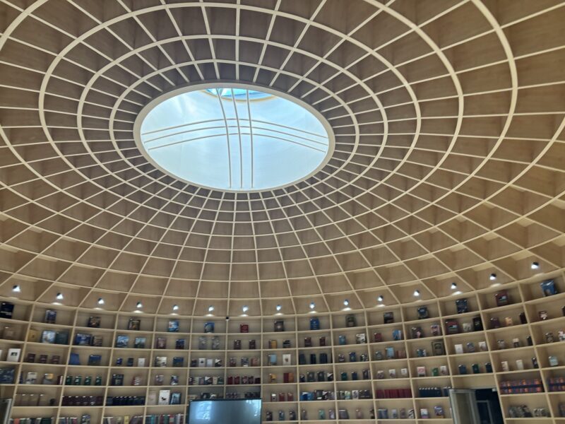 Low-angle shot of a library's dome-shaped wooden-supported ceiling and circular built-in bookshelves. The composition emphasizes the dome's concentric latticework leading to an oval skylight. Dominant warm brown tones are contrasted by natural light filtering through the skylight. Rows of books create a textured visual along the lower edge of the frame. Light is balanced throughout the image with the exception of the skylight which is overexposed and mostly white.