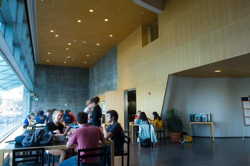 A dining area with tall ceilings and an empty wood wall. Students are sitting and eating.