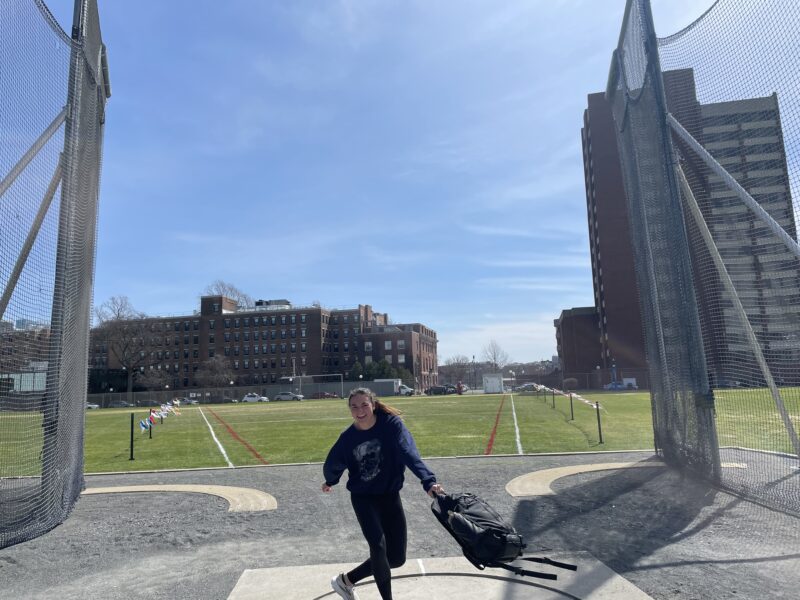 Girl doing shotput with her backpack in a green lane of grass.